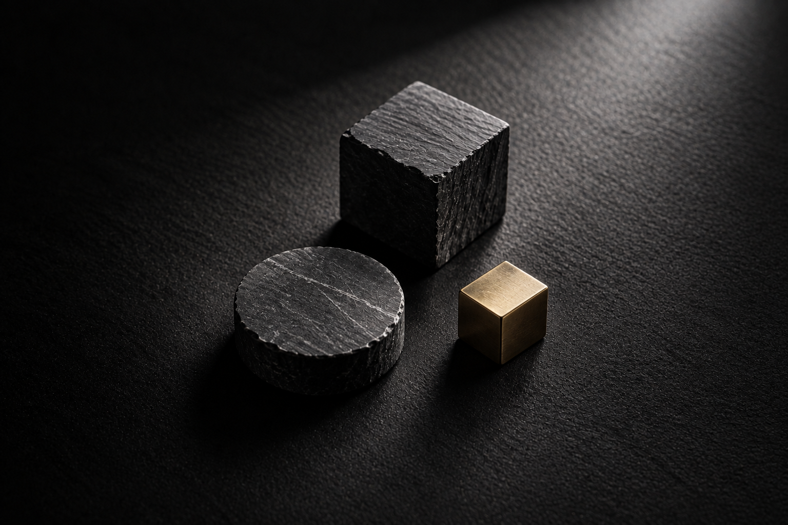 A dark stone block, a round stone form, and a brass cube centered on a black tabletop